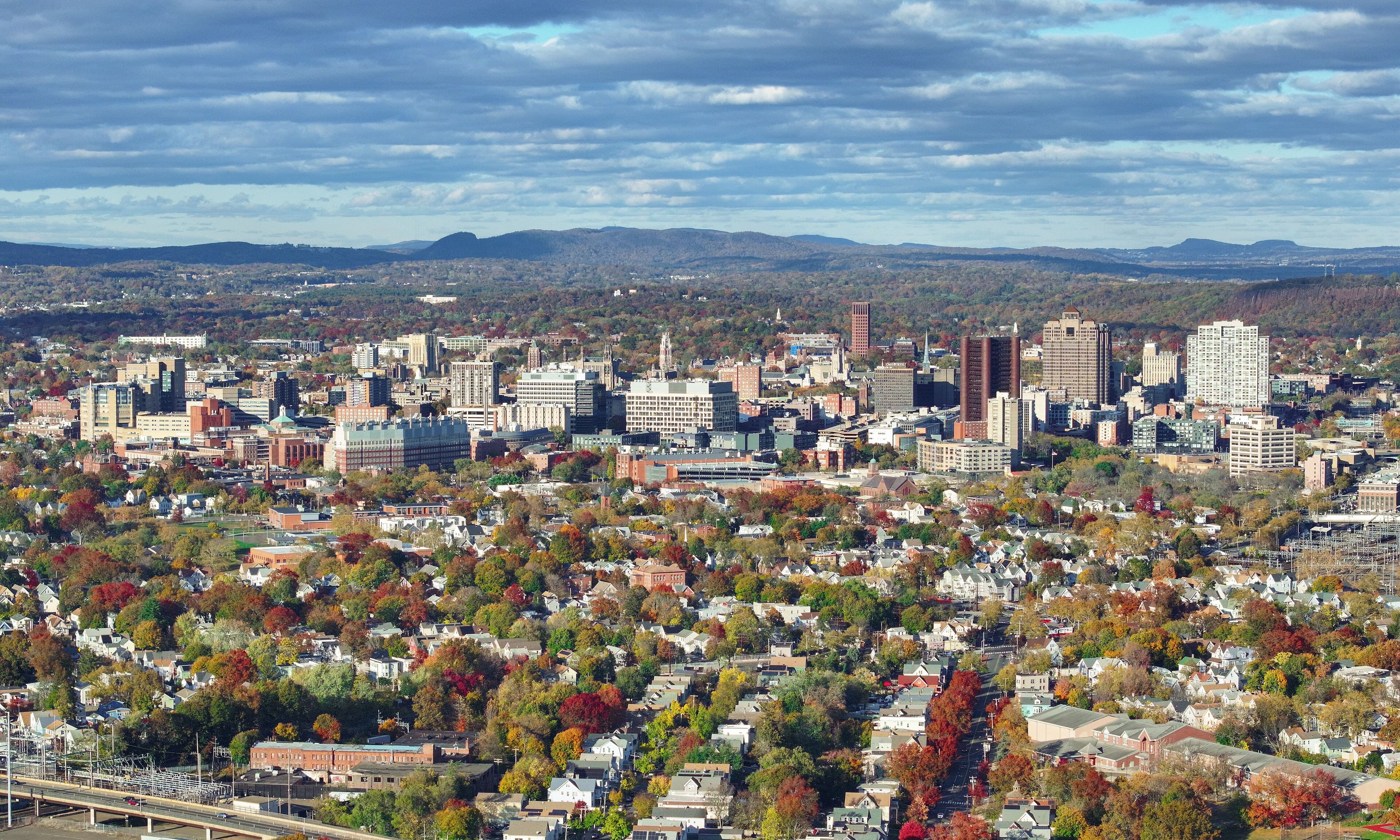 High-angle view of a city skyline surrounded by vibrant autumn trees and distant mountains.