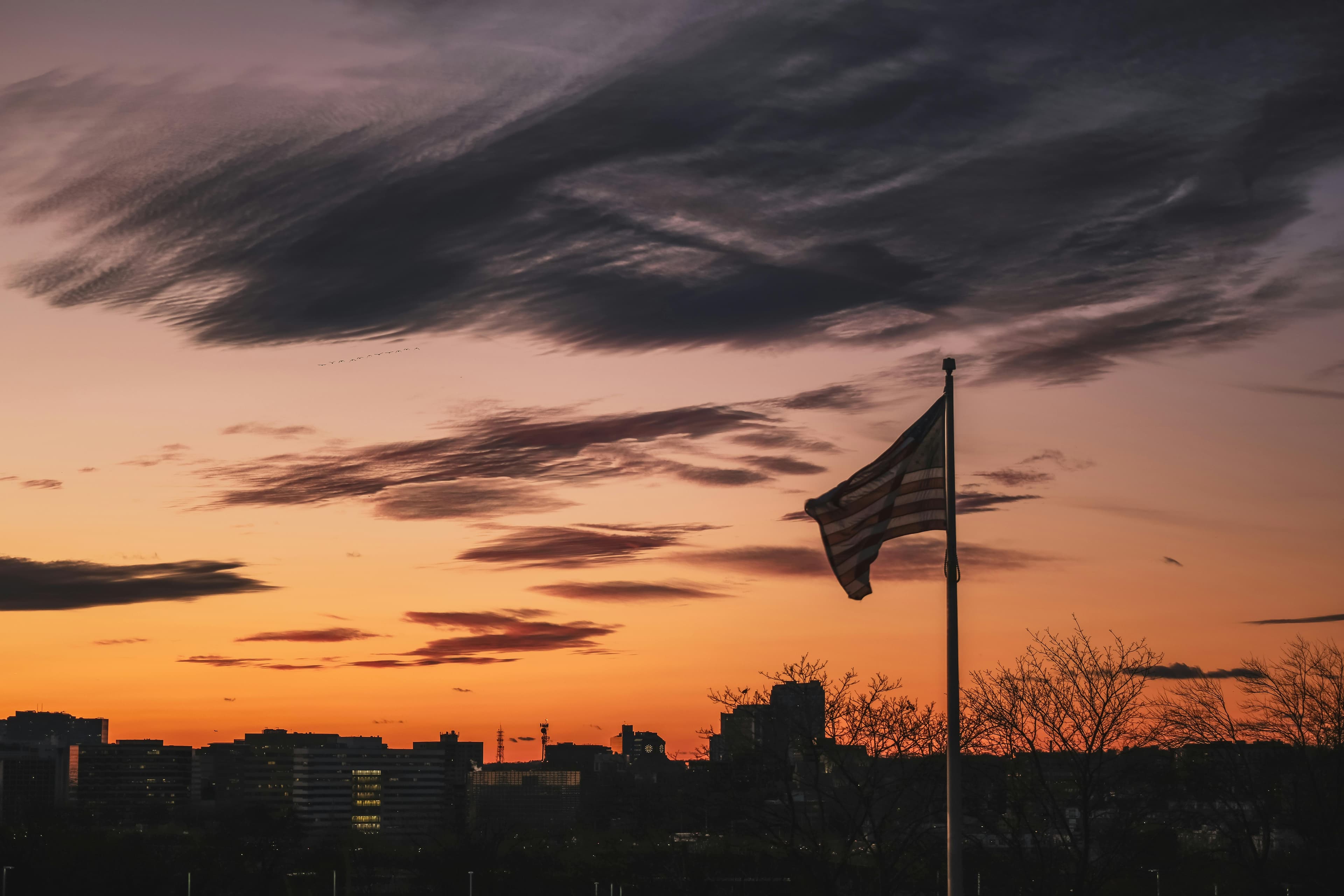 Silhouetted American flag against a vibrant orange and purple sunset sky over a city skyline.
